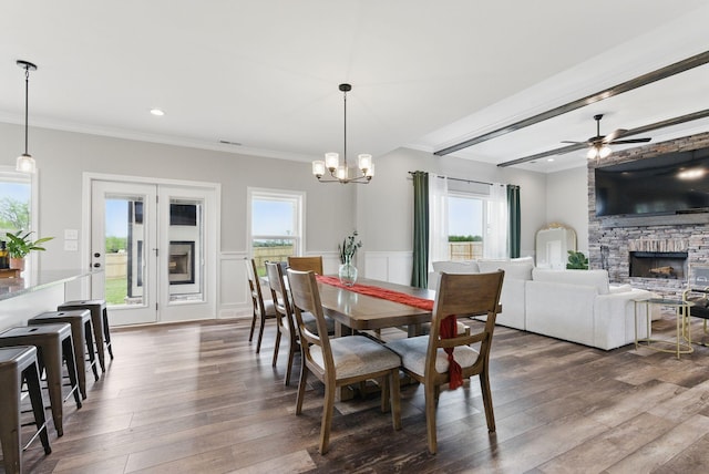 dining space featuring ornamental molding, ceiling fan with notable chandelier, a stone fireplace, and dark wood-style flooring