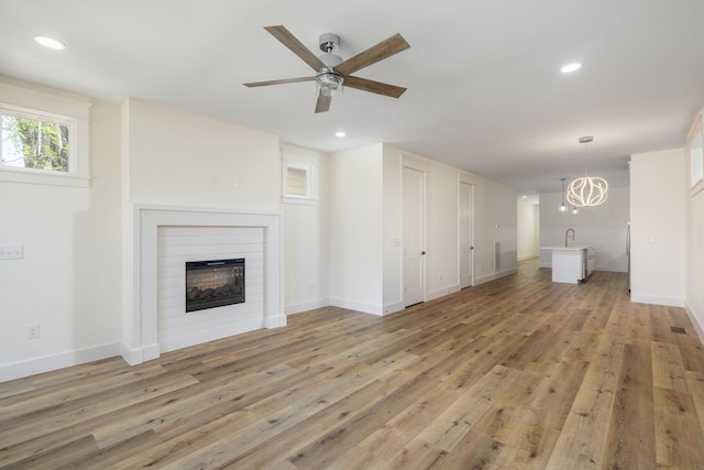 unfurnished living room featuring light wood-style floors, ceiling fan with notable chandelier, a glass covered fireplace, recessed lighting, and a sink