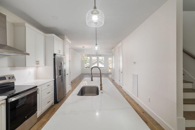 kitchen with wall chimney range hood, appliances with stainless steel finishes, a sink, decorative light fixtures, and light wood finished floors