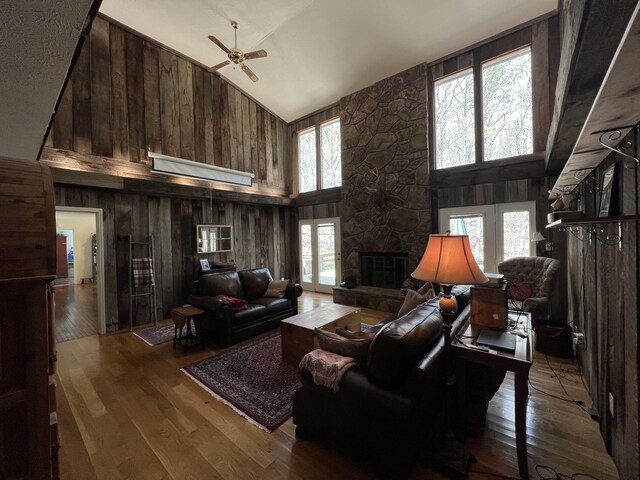 living room featuring high vaulted ceiling, a ceiling fan, wood finished floors, a stone fireplace, and wood walls