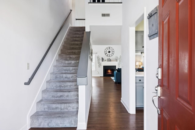 entryway with visible vents, a warm lit fireplace, dark wood-style floors, a towering ceiling, and stairs