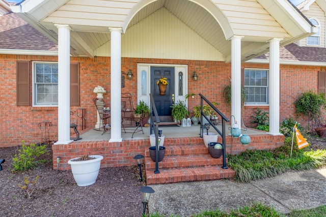 view of exterior entry with a porch, a shingled roof, and brick siding