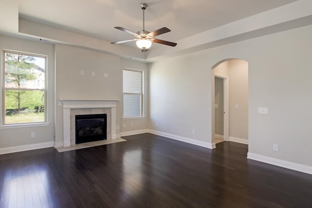 unfurnished living room with arched walkways, a tray ceiling, a ceiling fan, and wood finished floors