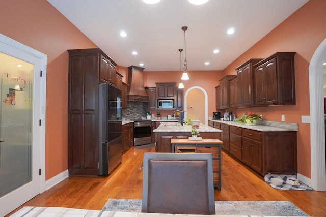 kitchen featuring an island with sink, arched walkways, stainless steel appliances, custom range hood, and a sink
