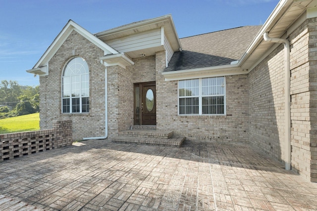 entrance to property featuring brick siding, a patio area, and roof with shingles