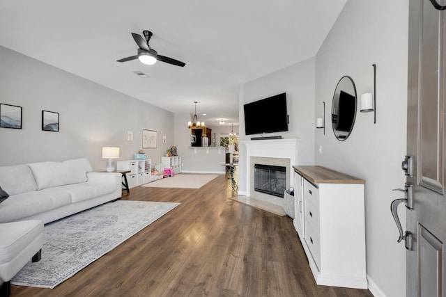 living area with a fireplace with flush hearth, visible vents, baseboards, dark wood-style flooring, and ceiling fan with notable chandelier