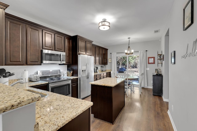 kitchen with an inviting chandelier, dark brown cabinets, light stone countertops, a kitchen island, and appliances with stainless steel finishes
