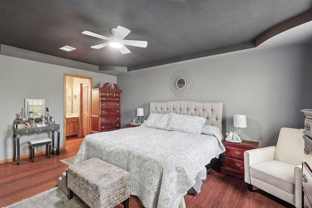 bedroom featuring a tray ceiling, wood finished floors, visible vents, connected bathroom, and ceiling fan
