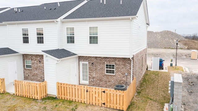 view of property exterior featuring brick siding, fence, roof with shingles, and central air condition unit
