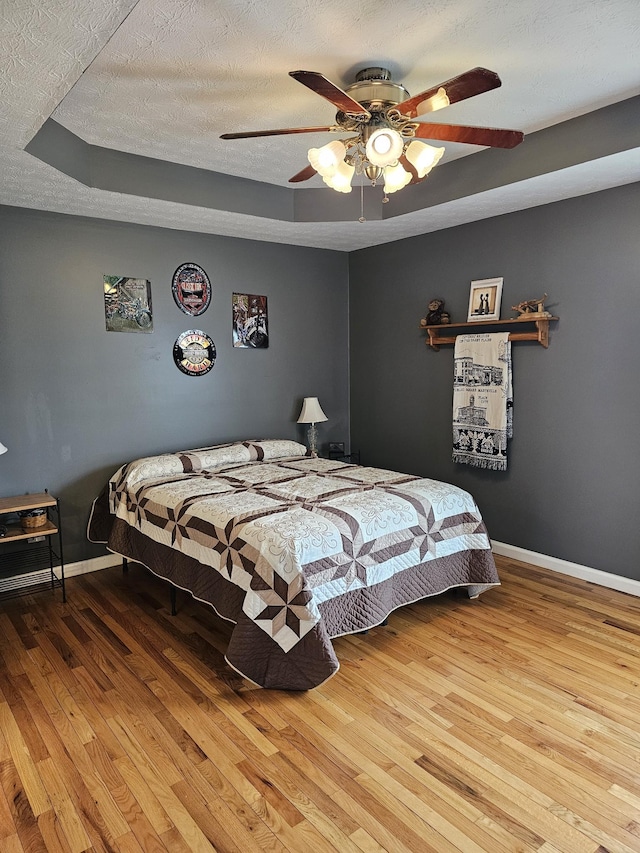 bedroom with a textured ceiling, baseboards, wood finished floors, ceiling fan, and a raised ceiling