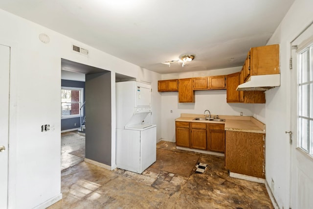 kitchen with a sink, baseboards, stacked washer / dryer, brown cabinetry, and under cabinet range hood