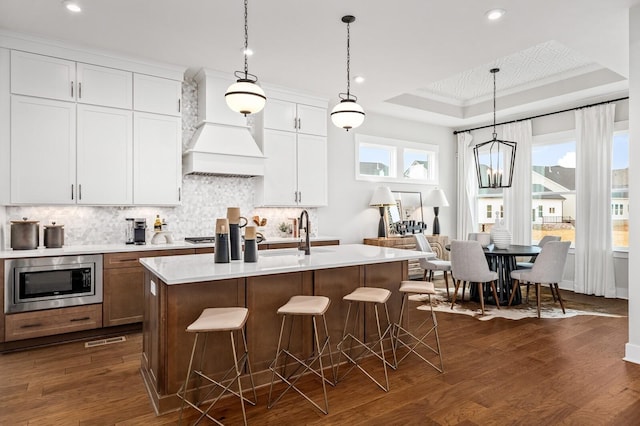 kitchen with stainless steel microwave, custom exhaust hood, backsplash, light countertops, and a raised ceiling