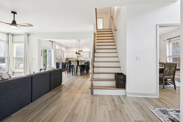 living area with a ceiling fan, baseboards, stairway, and light wood-type flooring