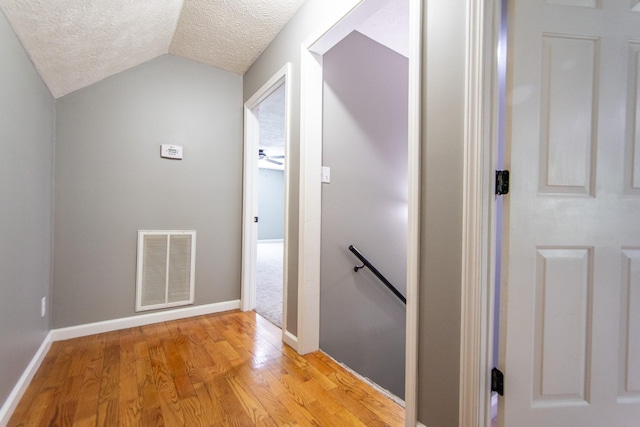 corridor with vaulted ceiling, an upstairs landing, a textured ceiling, light wood-type flooring, and visible vents