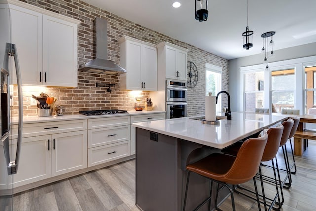 kitchen with a sink, white cabinetry, stainless steel double oven, wall chimney range hood, and light countertops
