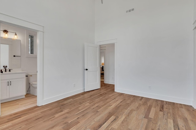 unfurnished bedroom with light wood finished floors, baseboards, visible vents, a high ceiling, and ensuite bath