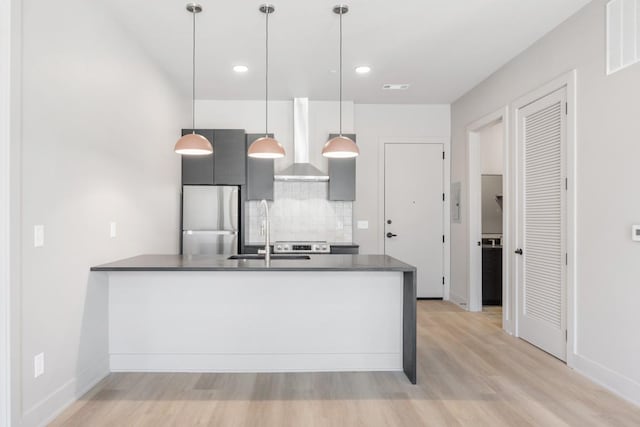 kitchen featuring freestanding refrigerator, wall chimney range hood, dark countertops, light wood-style flooring, and a peninsula