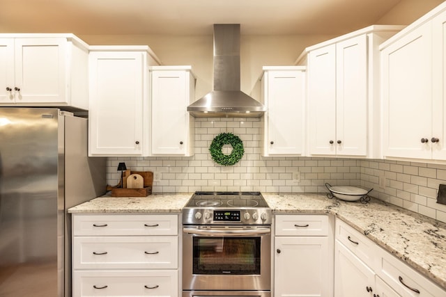kitchen with white cabinets, backsplash, light stone counters, wall chimney range hood, and appliances with stainless steel finishes