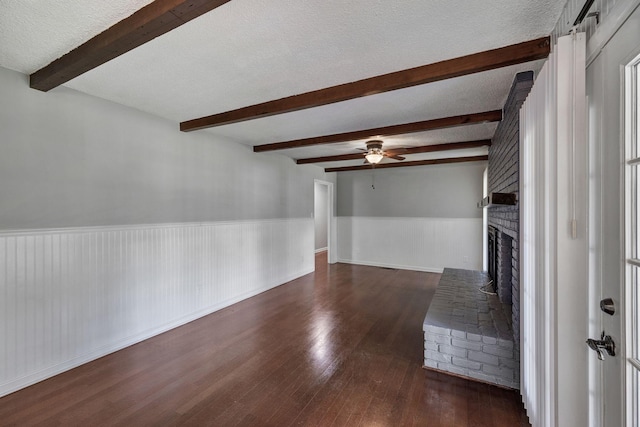 unfurnished living room featuring beam ceiling, dark wood-style flooring, wainscoting, a ceiling fan, and a fireplace