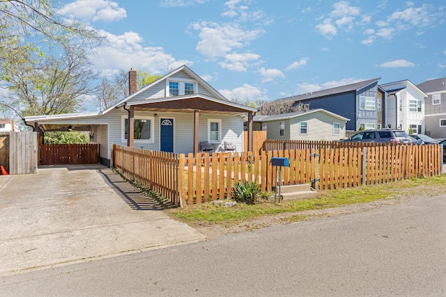 bungalow-style home featuring a fenced front yard, a chimney, concrete driveway, a carport, and covered porch