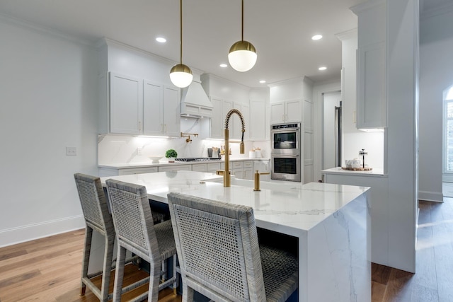 kitchen with light wood finished floors, crown molding, a kitchen island with sink, appliances with stainless steel finishes, and white cabinetry