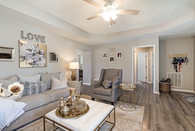 living room with a tray ceiling, visible vents, baseboards, wood finished floors, and ceiling fan