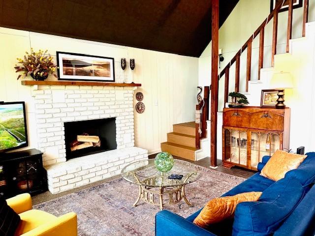 carpeted living room featuring vaulted ceiling, a brick fireplace, and stairway