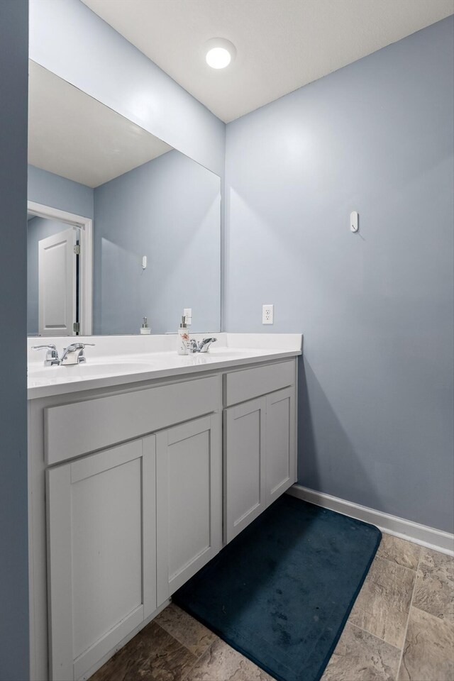 bathroom featuring baseboards, stone tile flooring, a sink, and double vanity