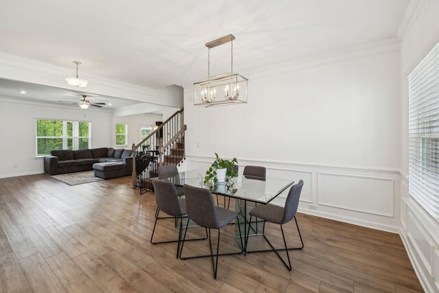 dining room featuring crown molding, wood finished floors, stairs, ceiling fan with notable chandelier, and a wainscoted wall