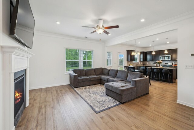 living area with light wood finished floors, ornamental molding, baseboards, a glass covered fireplace, and ceiling fan