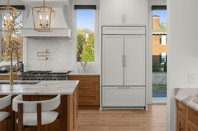 kitchen featuring built in refrigerator, tasteful backsplash, light wood-style floors, custom exhaust hood, and a notable chandelier