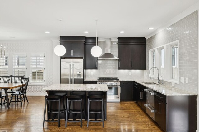 kitchen featuring wall chimney range hood, pendant lighting, a sink, appliances with stainless steel finishes, and a center island