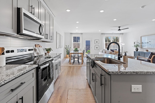 kitchen with a sink, stainless steel appliances, light wood-type flooring, light stone countertops, and an island with sink