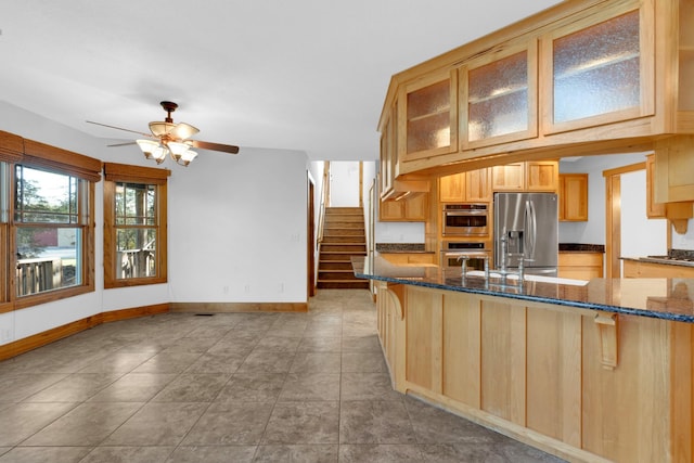 kitchen featuring dark stone countertops, glass insert cabinets, light brown cabinets, stainless steel appliances, and ceiling fan