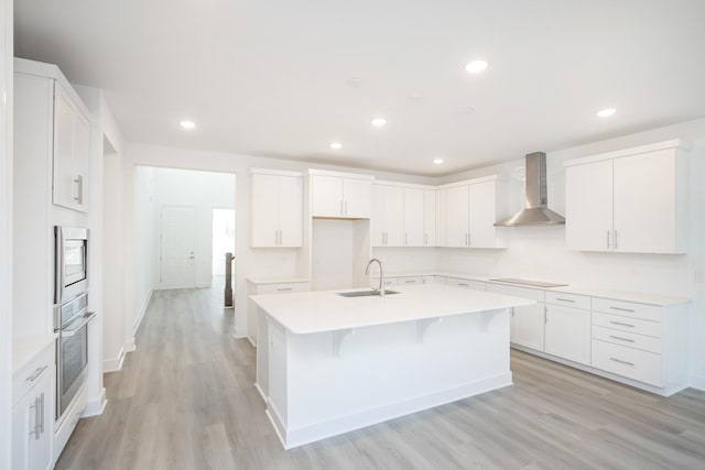 kitchen featuring oven, black electric cooktop, a sink, wall chimney exhaust hood, and a center island with sink