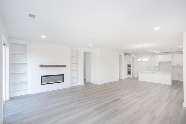 unfurnished living room featuring recessed lighting, light wood finished floors, a notable chandelier, built in features, and a glass covered fireplace