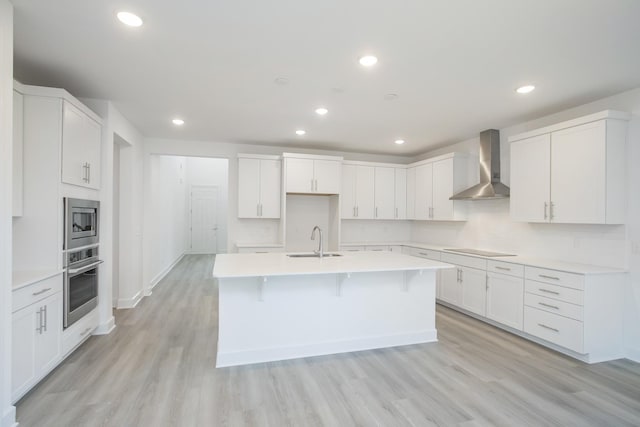 kitchen featuring a kitchen island with sink, recessed lighting, a sink, wall chimney range hood, and appliances with stainless steel finishes