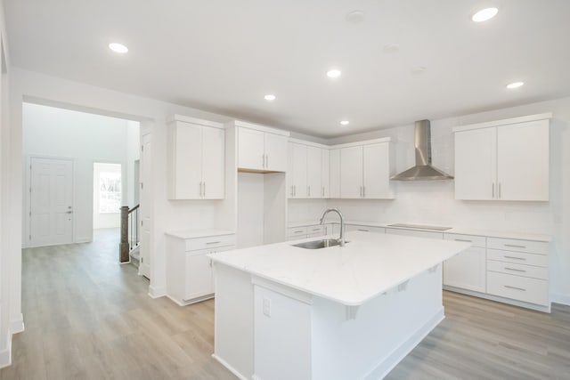 kitchen featuring light wood finished floors, a kitchen island with sink, a sink, white cabinetry, and wall chimney range hood