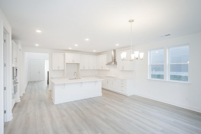 kitchen with wall chimney range hood, a sink, a chandelier, light wood finished floors, and an island with sink