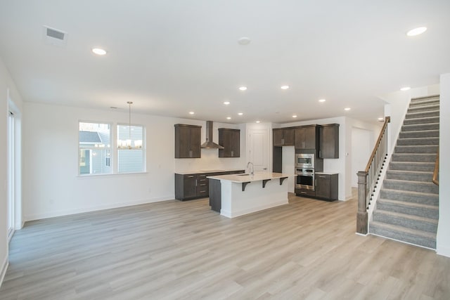 kitchen with a sink, light wood-type flooring, wall chimney exhaust hood, dark brown cabinetry, and visible vents