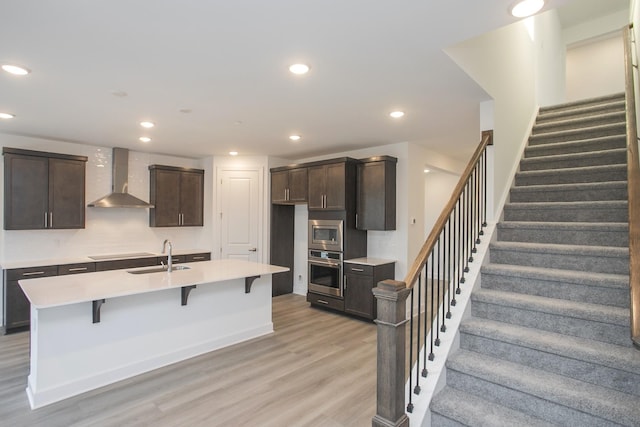 kitchen with stainless steel appliances, wall chimney range hood, dark brown cabinets, light wood finished floors, and a sink