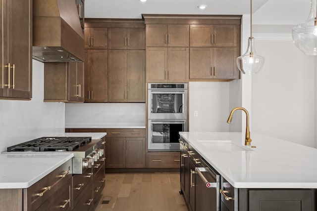 kitchen featuring custom range hood, a sink, stainless steel appliances, light wood finished floors, and a center island with sink