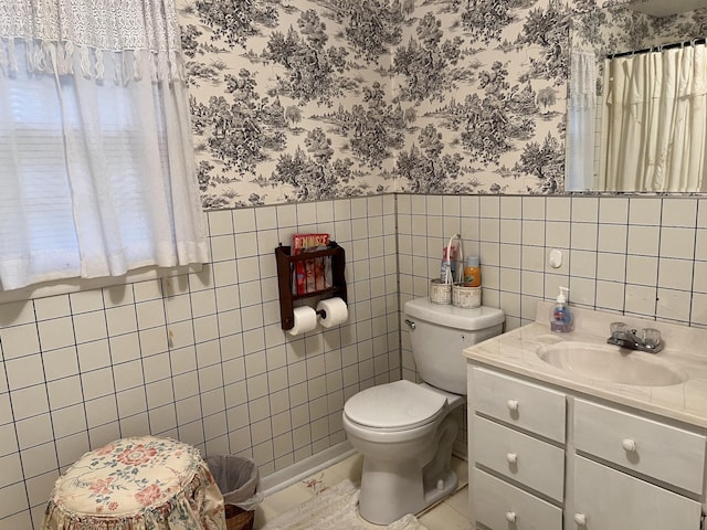 full bathroom with a wainscoted wall, wallpapered walls, toilet, tile walls, and vanity
