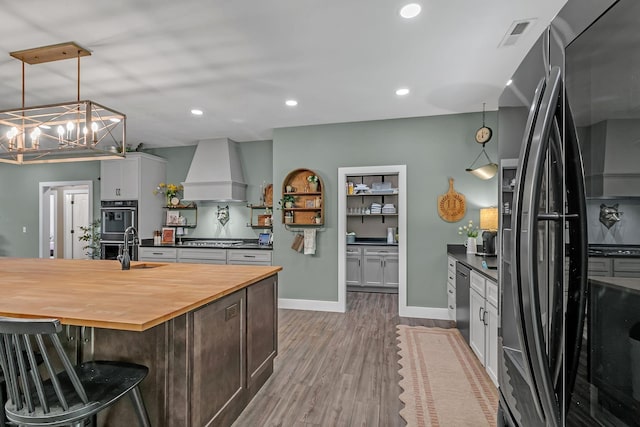 kitchen with butcher block counters, custom range hood, black appliances, visible vents, and wood finished floors
