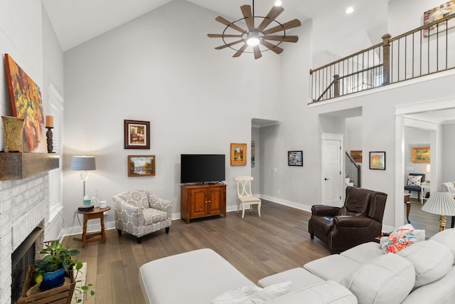 living area with dark wood-style flooring, a brick fireplace, high vaulted ceiling, stairs, and a ceiling fan