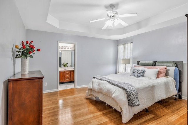 bedroom with baseboards, a tray ceiling, a ceiling fan, light wood-style flooring, and ensuite bath