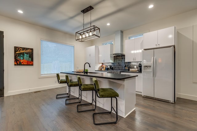 kitchen featuring wall chimney range hood, white fridge with ice dispenser, a sink, a kitchen bar, and a center island with sink