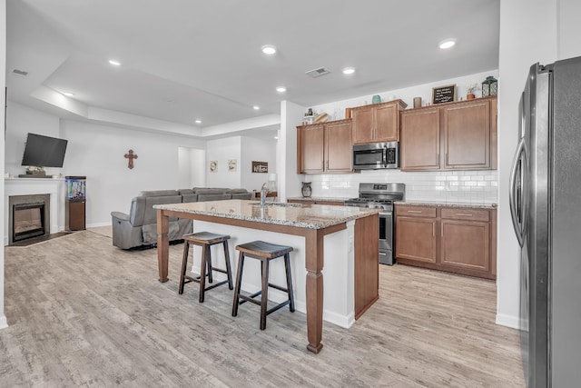 kitchen with appliances with stainless steel finishes, light stone countertops, light wood finished floors, open floor plan, and an island with sink