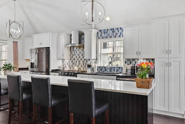 kitchen with a sink, stainless steel appliances, a notable chandelier, tasteful backsplash, and wall chimney exhaust hood