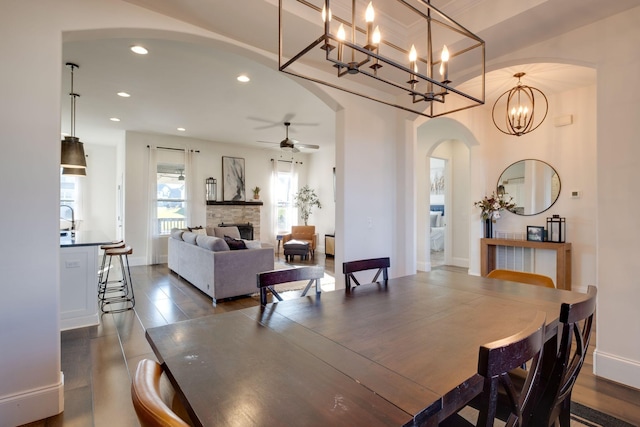 dining room with arched walkways, recessed lighting, dark wood-type flooring, a stone fireplace, and ceiling fan with notable chandelier